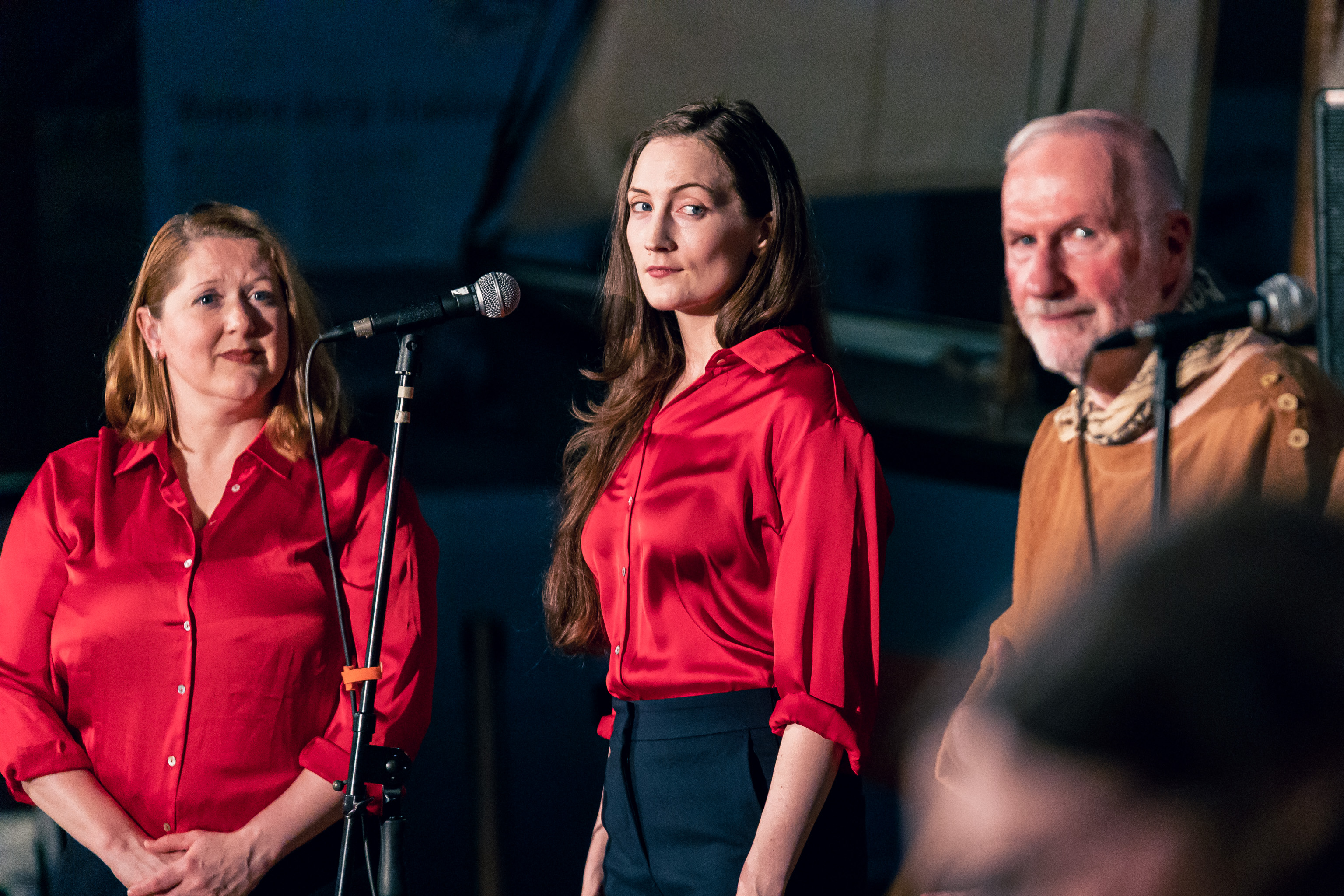 Elizabeth Morris, Lesley Smith, Jim McDermott look over their left shoulder, toward the camera. Elizabeth and Lesley are wearing red shirts, Jim is wearing a brown shirt.