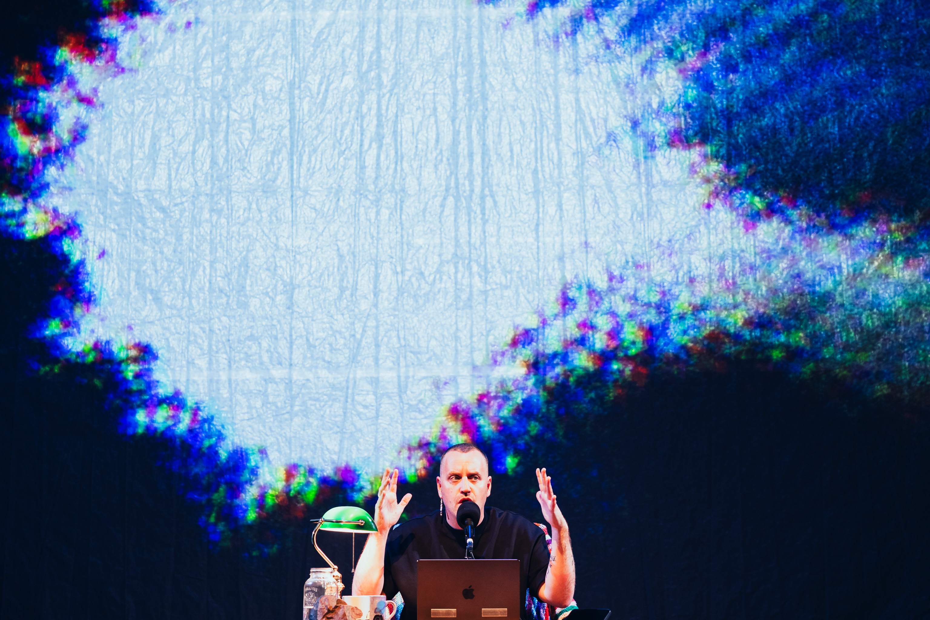 Stewart Legere, seated at a table with a computer infant of him. He hold his hands up beside his head.