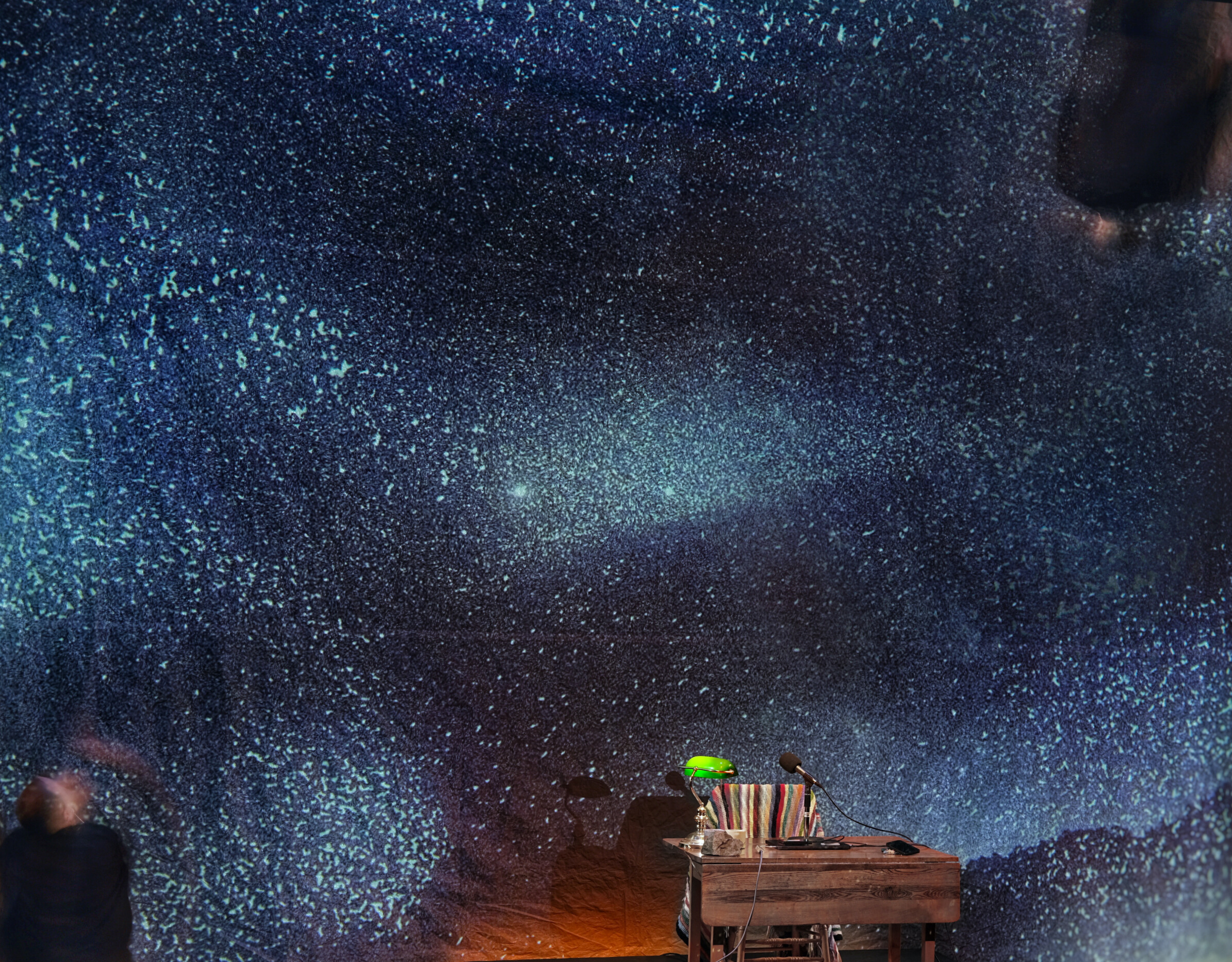 a table with a computer and green banker's light. a backdrop with projected starry sky. a man in black exits the left of the frame.