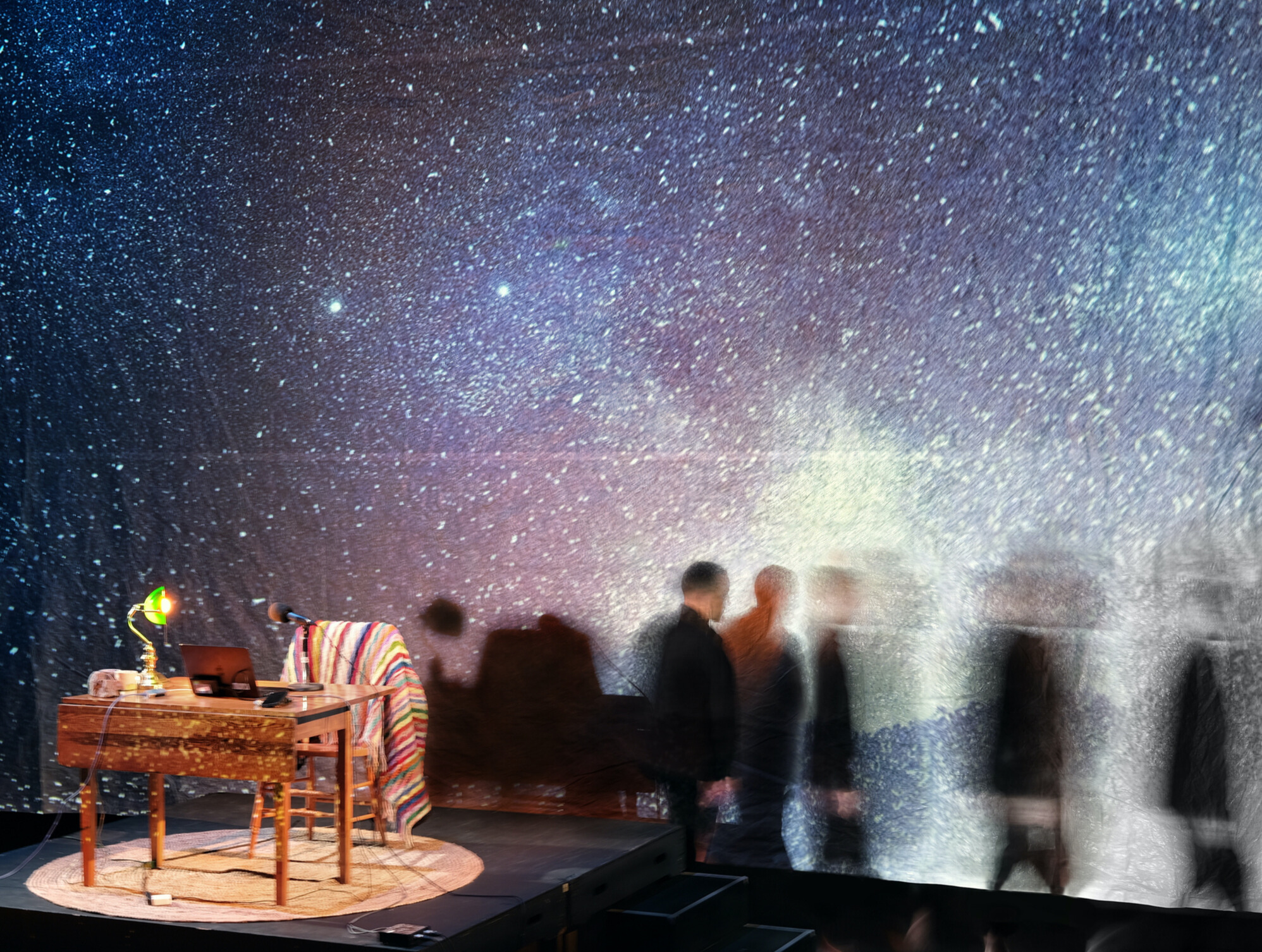 A giant projection screen with an image of the night sky, dotted with millions of stars, in blue, and purple, and black. On the left of the image, a small wooden table, with a green banker's light, a small grey rock, and an open laptop. A cozy wooden armchair is pulled up to the table, with a colourful knit blanket draped over the back. These things all sit atop a circular beige jute rug. A man, dressed in black, walks away from the table. The image is over exposed, so ghostly reflections of the man cascade out of frame.  