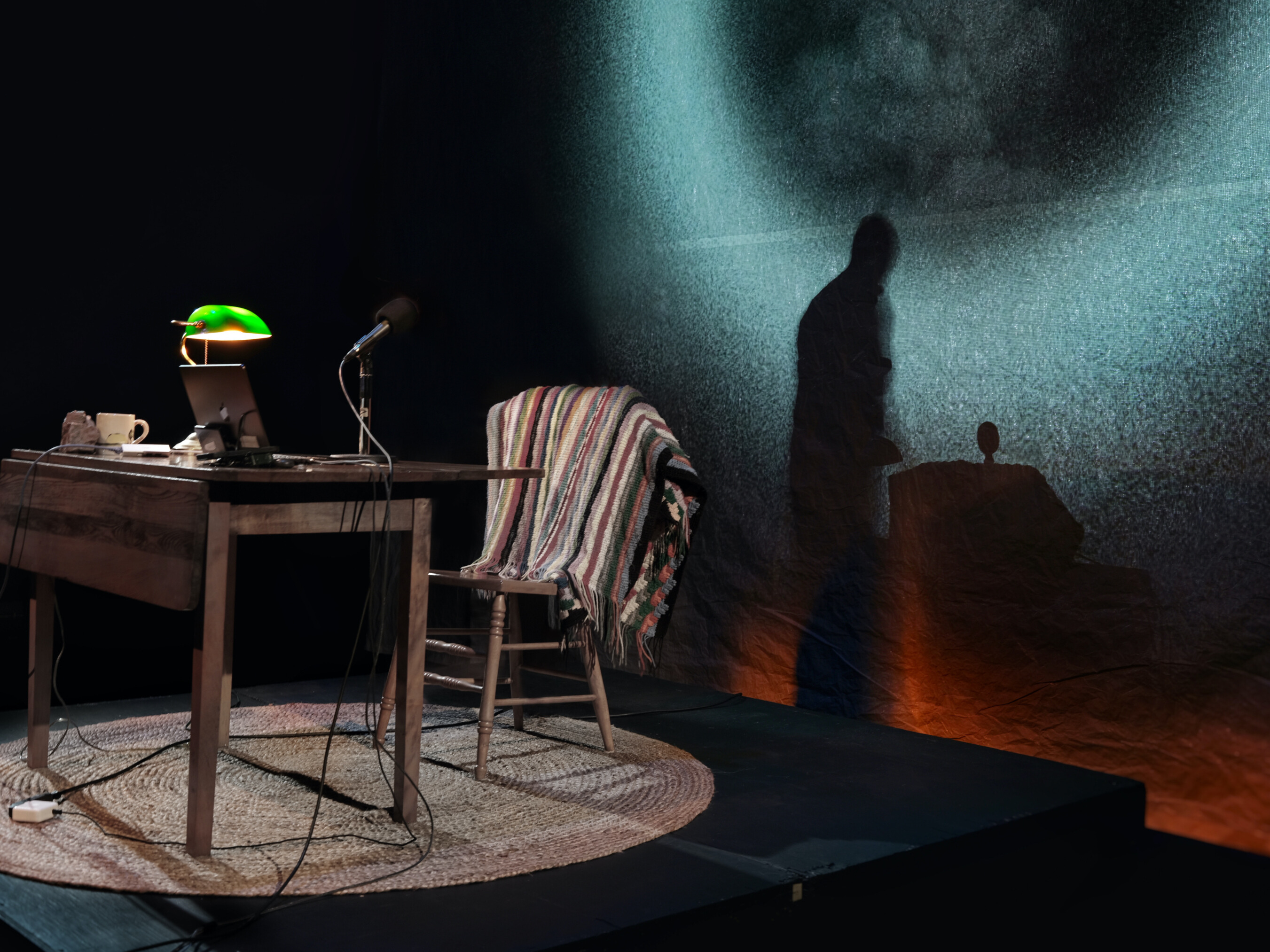to the left, a chair with a blanket, a table, a banker's lamp. behind it, a large glowing blue circle on the wall. a shadow of a man on the wall. 