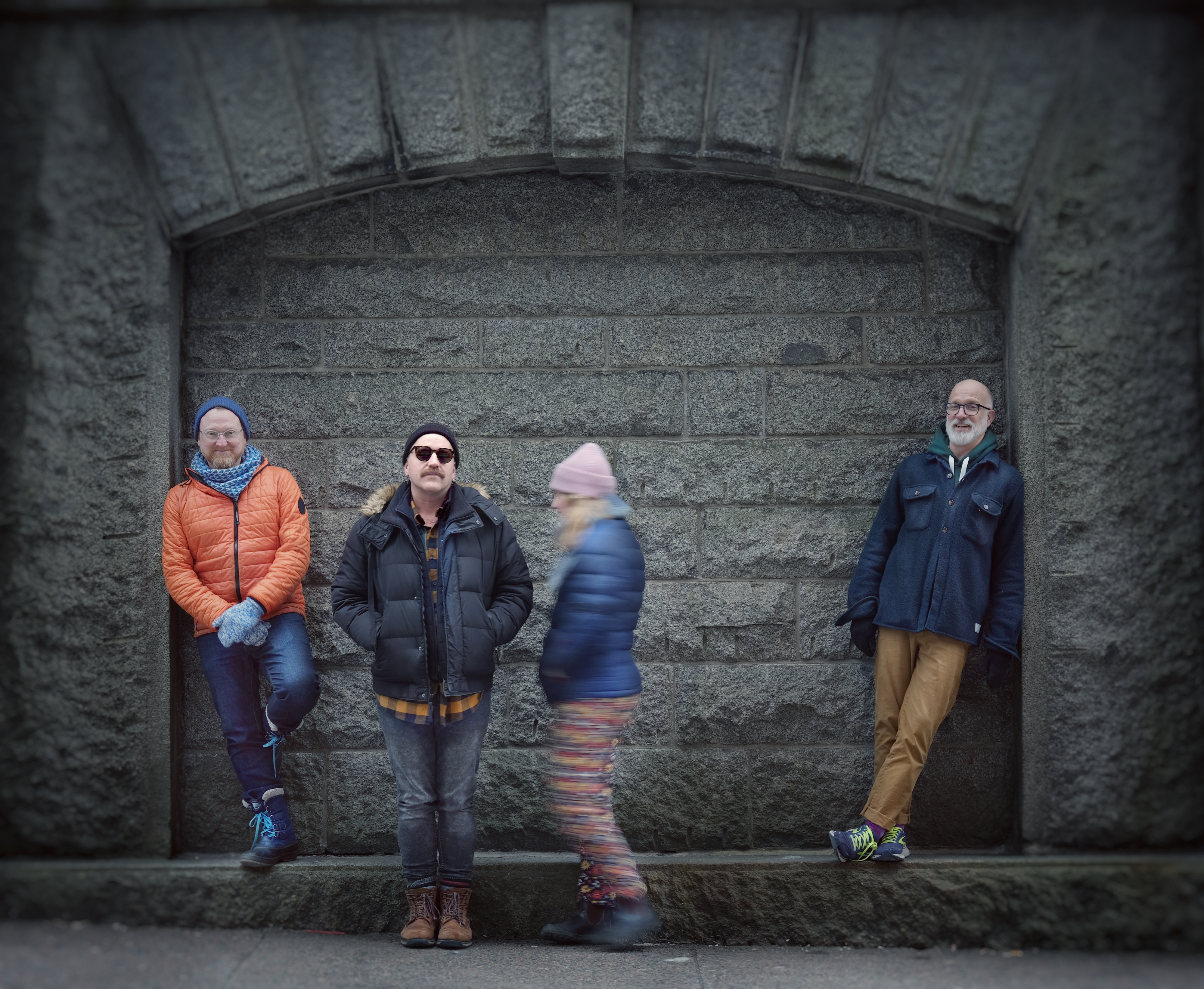 A grey stone wall background with a shallow wall well. L to R: Alex is leaning agains the right side of wall well, looking forward. Stewart is standing a few feet forward, with his hands in his pockets. He is wearing sunglasses and looking forward. Sophie stands next to him, facing him. She wears a pink hat and is moving, so her image is slightly blurred. Ben is leaning against the left side of the wall well.