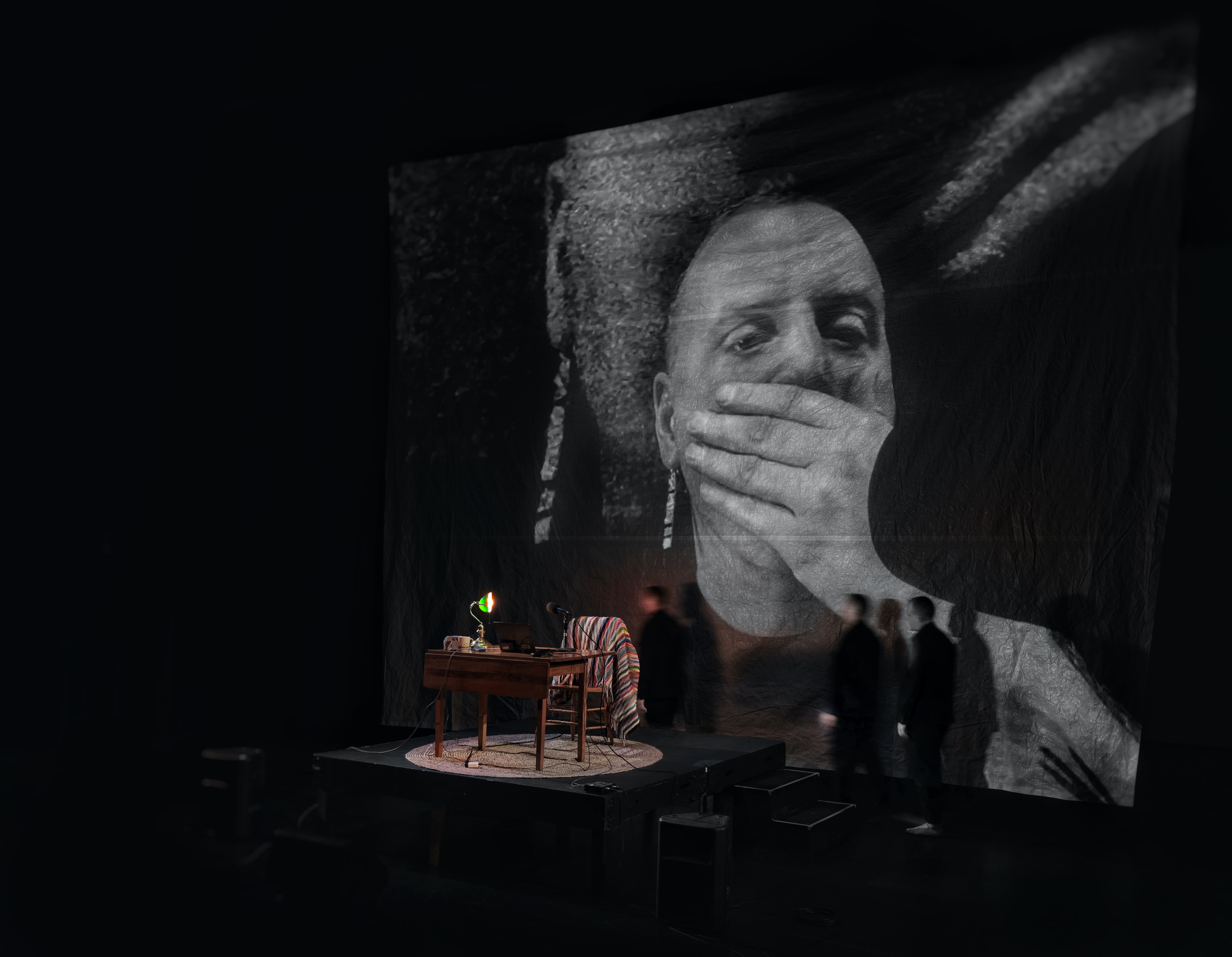 a table with a green banker's light. a chair with a rainbow blanket draped over the back. a man sits at the chair. his face is projected on the wall, very large, behind him. he is covering his mouth, as if in shock. 