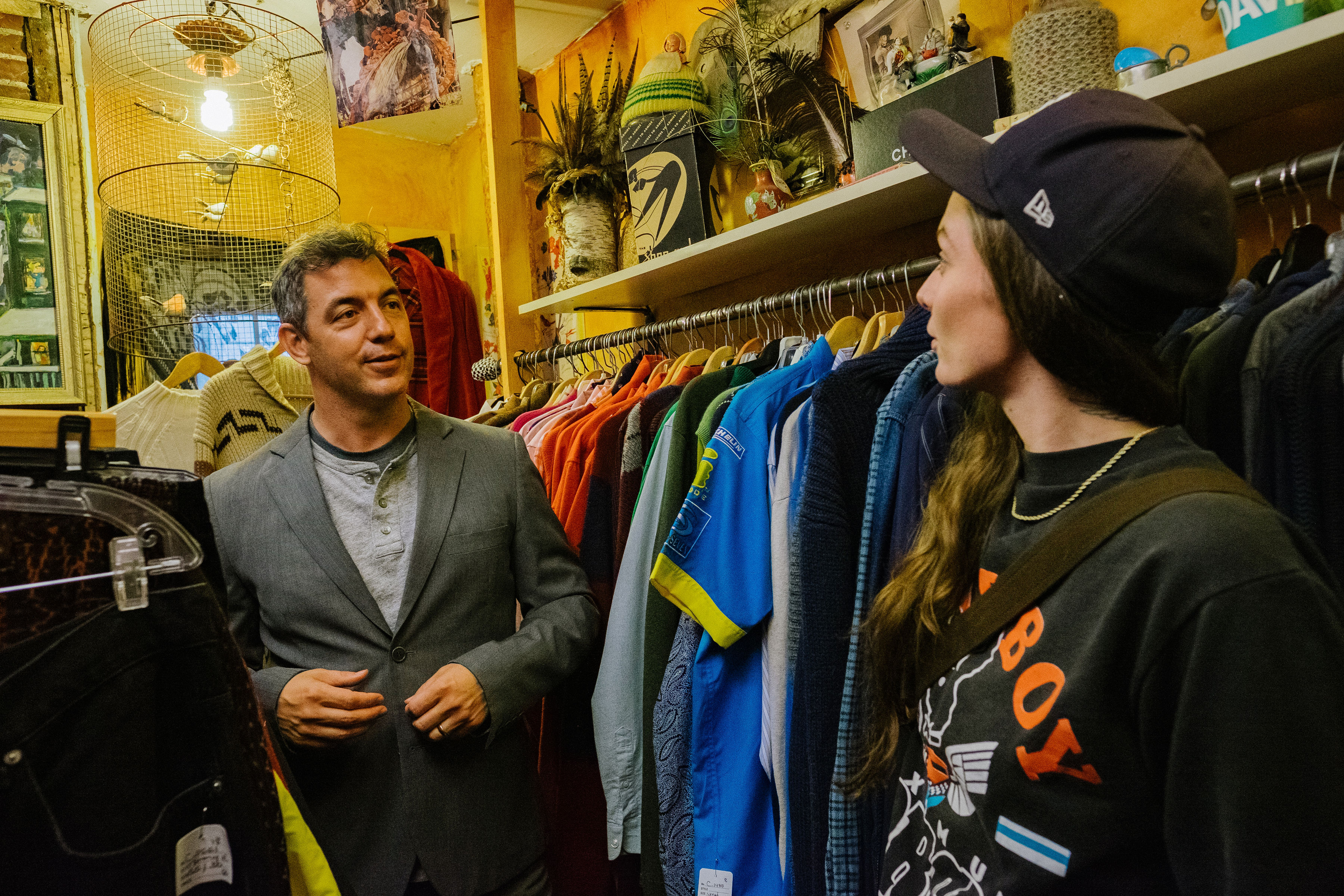 A man talks to an audience member in the back of a used clothing store.