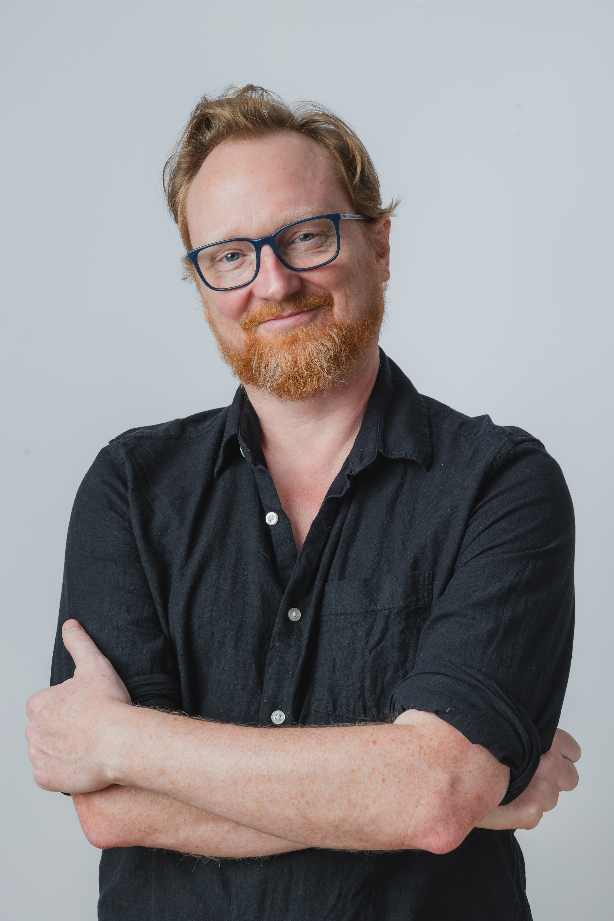 A picture of Alex McLean. He is wearing a blue button-up shirt. He is standing with his arms crossed, his head slightly cocked to one side, and has a pleasant smile on his face. The background is white. 
