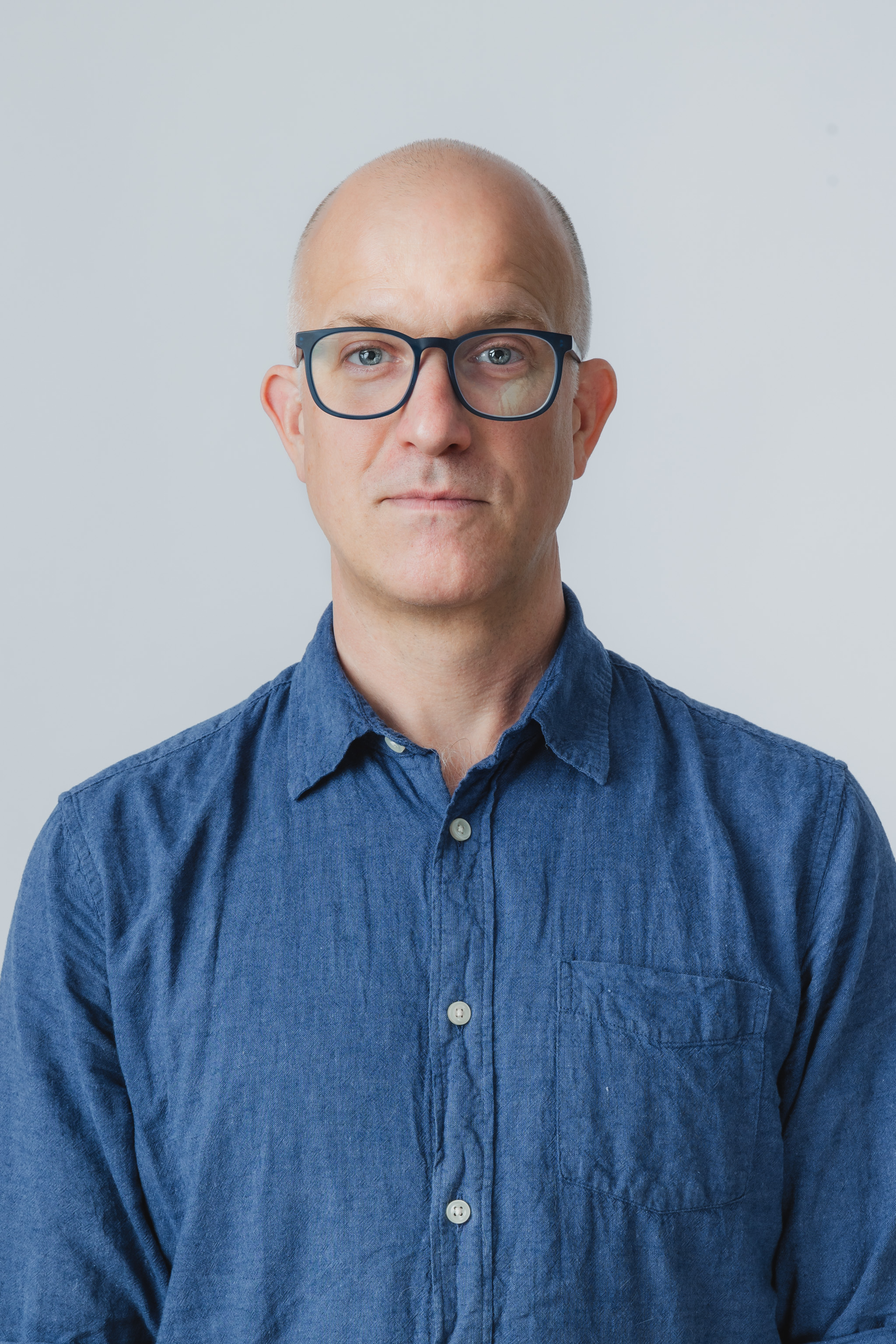 A picture of Ben Stone. From mid torso up. He is wearing a light blue button up shirt. He is squared perfectly to the camera. He has a neutral expression on his face, with the slightest hint of a smile in his eyes.  