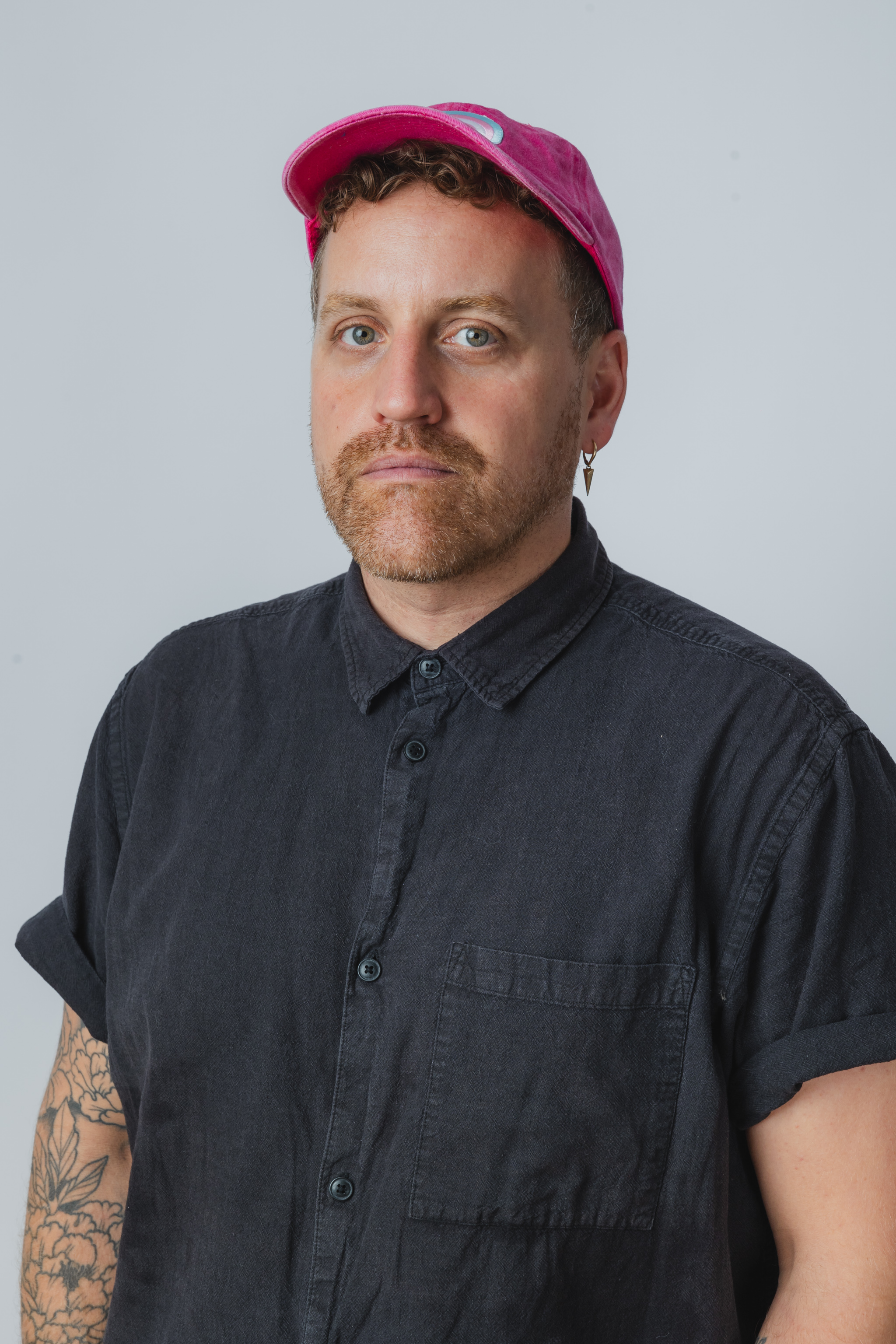 A picture of Stewart Legere. He is slightly angled, looking at the camera. He is wearing a dark blue short-sleeved button up shirt. He is wearing a pink ballcap with a Trans flag on the front. He has short, reddish stubble, and a neutral expression that indicates he doesn't love having his picture taken. 